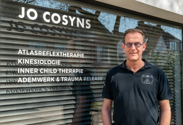 Jo Cosyns bij de ingang van zijn praktijk. Op het raam van de gevel naast zich staan de naam van zijn praktijk en zijn specialisaties.